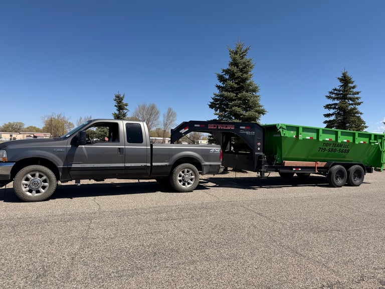 Silver pickup truck towing a black gooseneck trailer and green dump trailer in a parking lot with evergreen trees in background