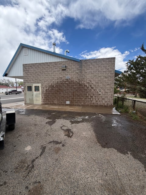 Brown brick building with white roof section under blue sky with clouds, gravel lot, and mountains in background