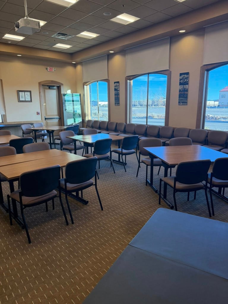 Modern office meeting room with multiple tables, black chairs, large windows overlooking water, and contemporary ceiling lights