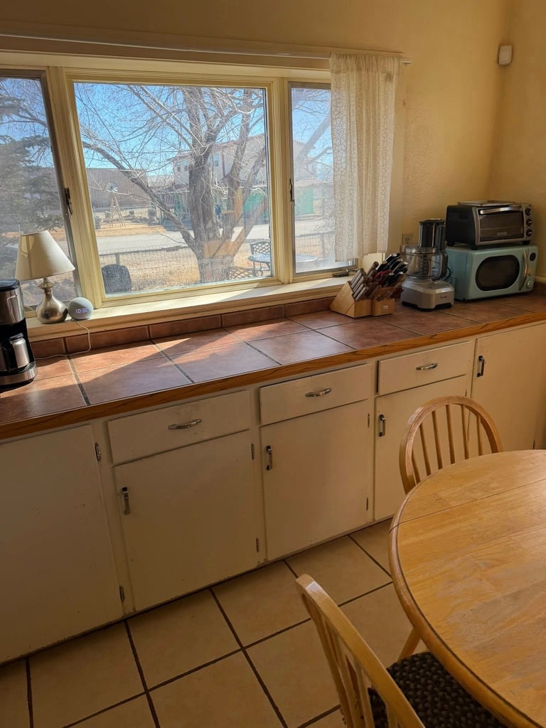 Vintage kitchen with cream cabinets, wooden countertop, large windows overlooking bare tree and outdoor landscape, wooden table in foreground