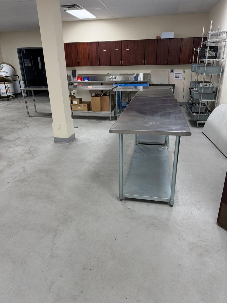 Commercial kitchen prep area with stainless steel work table, dark wood cabinets, concrete floor, and storage shelves in background