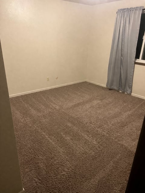 Empty room with beige walls, brown carpet, gray curtains covering a window, and electrical outlets on the wall