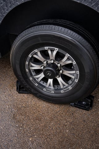 Vehicle wheel with two-tone silver and black alloy rim, mounted on asphalt with wheel chock visible underneath