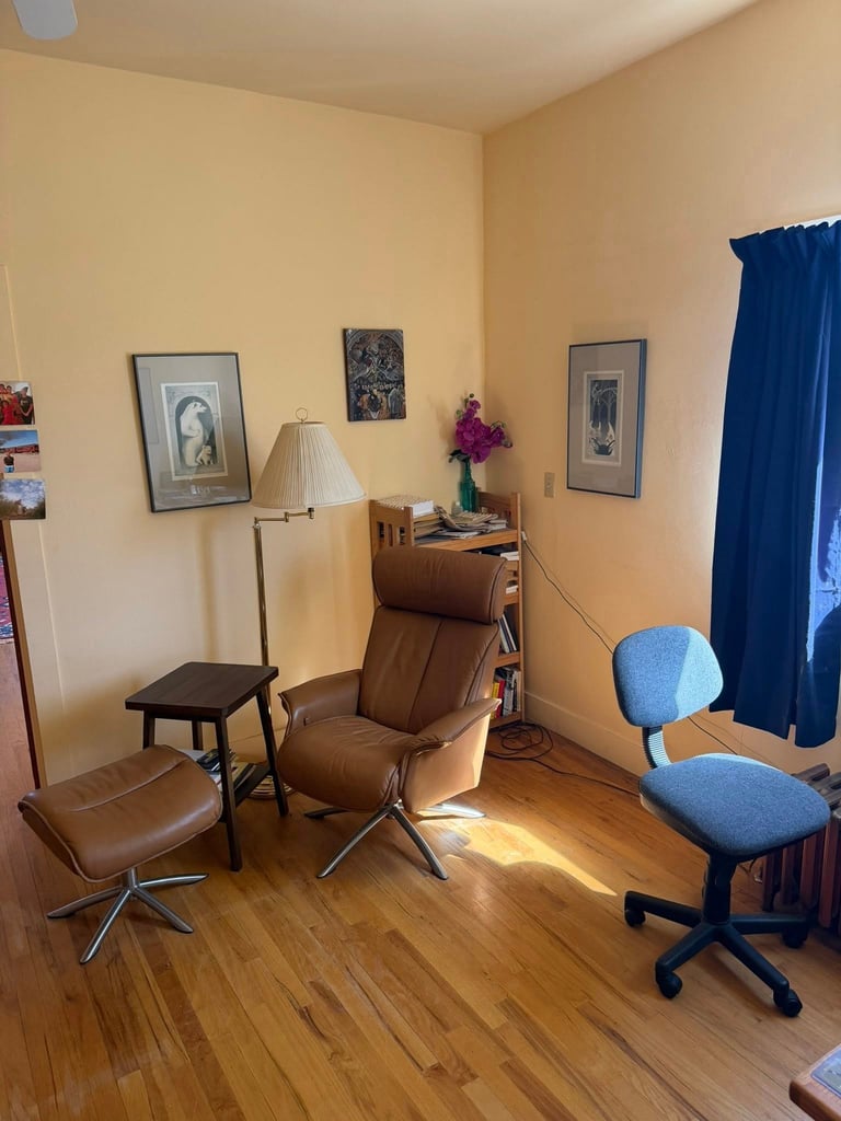 Cozy office room with beige walls, brown leather chair, blue desk chair, wooden desk, floor lamp, framed artwork, and blue curtains on hardwood floor