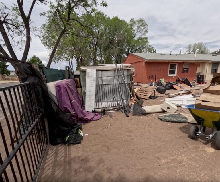 A cluttered residential yard with scattered materials, metal gates, a red building, and trees under an overcast sky