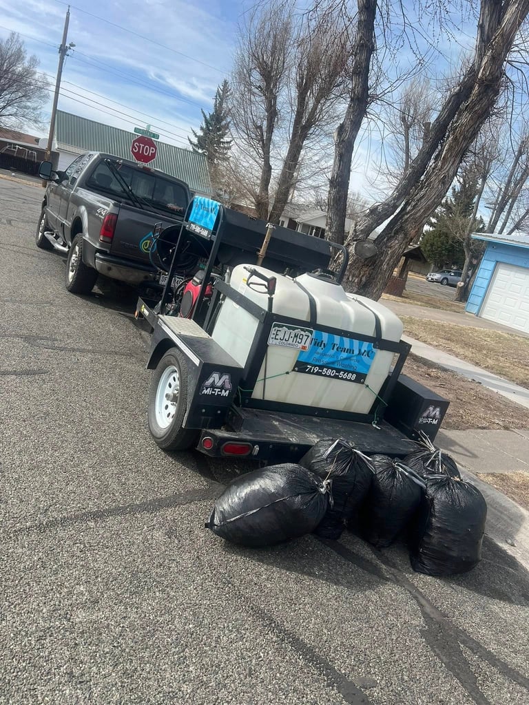 Small white and black equipment trailer loaded with black garbage bags parked in a lot with bare trees and vehicles in background