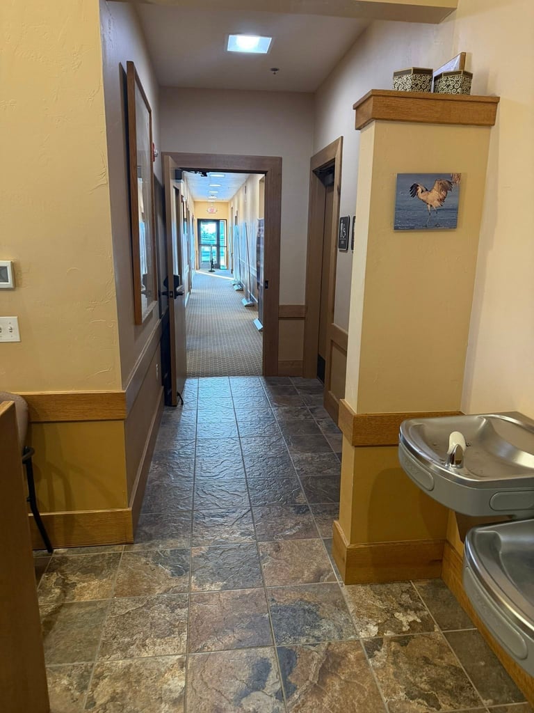 Long hallway with yellow walls, slate flooring, brown doors, water fountain, and artwork on walls with skylight ceiling