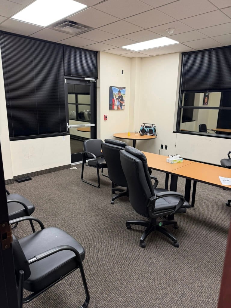 Modern office break room with black leather chairs, desks, mounted monitors on walls, and carpeted flooring under suspended ceiling