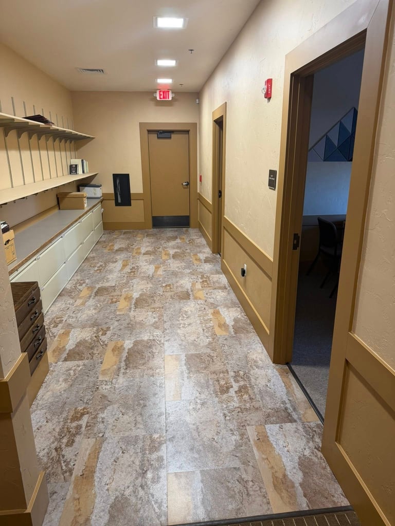 Modern office hallway with tile flooring, wooden doors, white walls, built-in shelving on left, and exit sign overhead