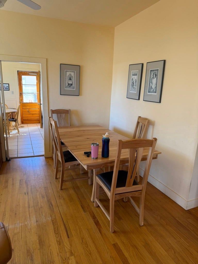Bright dining room with wooden table and chairs, cream yellow walls, framed artwork, and open doorway leading to another room