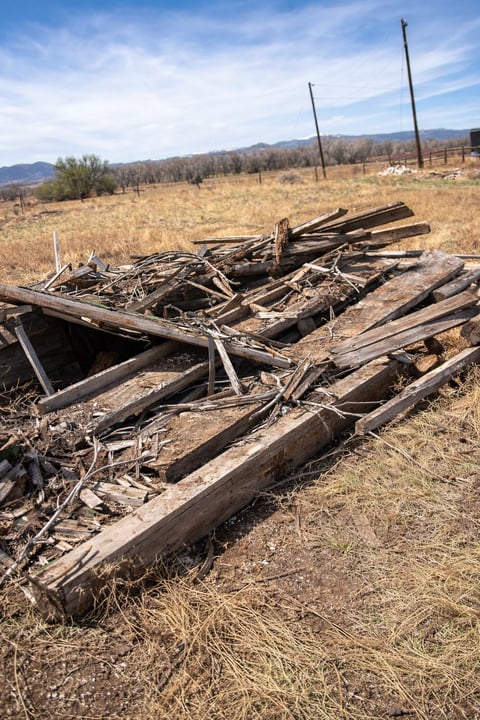 Pile of weathered wooden logs and timber scattered on dry, grassy terrain with mountains and power lines in the background