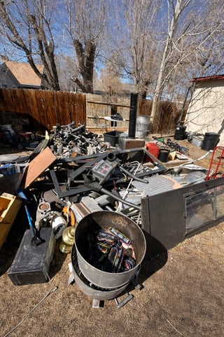 Junkyard scene with piles of scrap metal, old machinery, and discarded items in a residential yard with bare trees