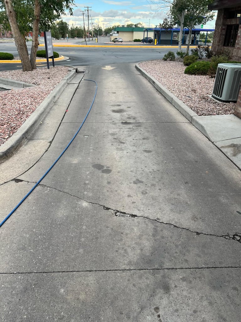 A concrete driveway with a blue hose running along the side, bordered by gravel and trees in a commercial parking area