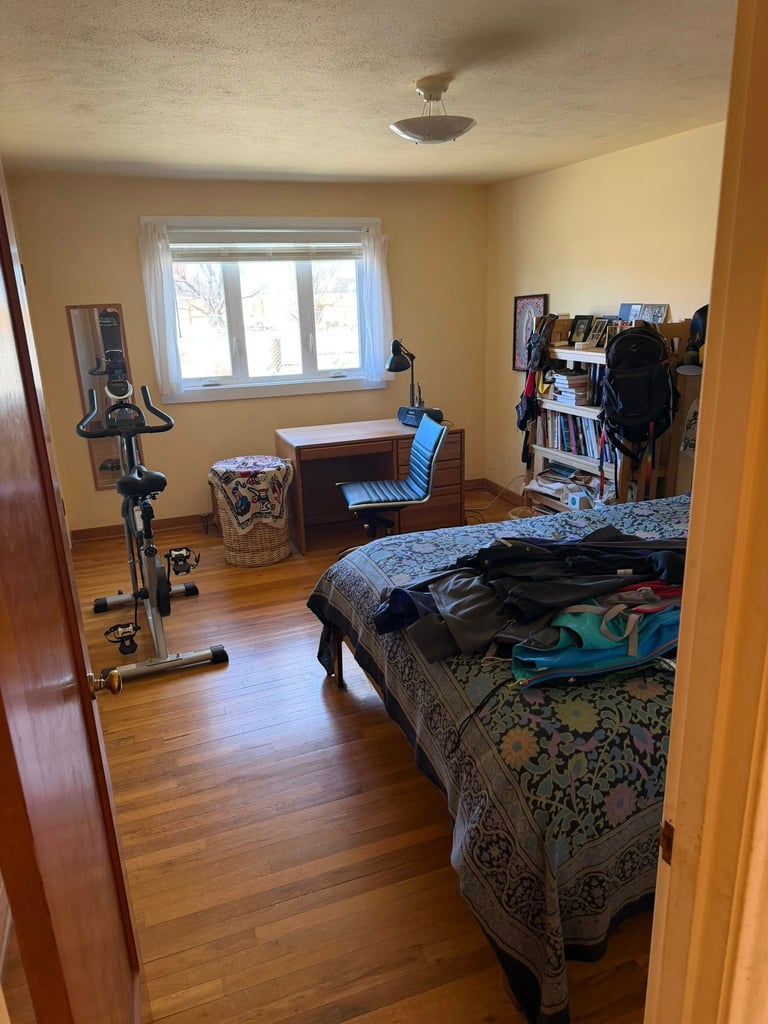 A spacious bedroom with yellow walls, wooden flooring, a bed with clothes on it, stationary bike, desk, and window with trees visible outside